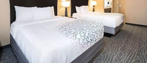 A hotel room with two beds, a large bed with a patterned bedspread and a smaller bed with a plain bedspread. There are two bedside lamps, a framed picture on the wall, and a door to the right.