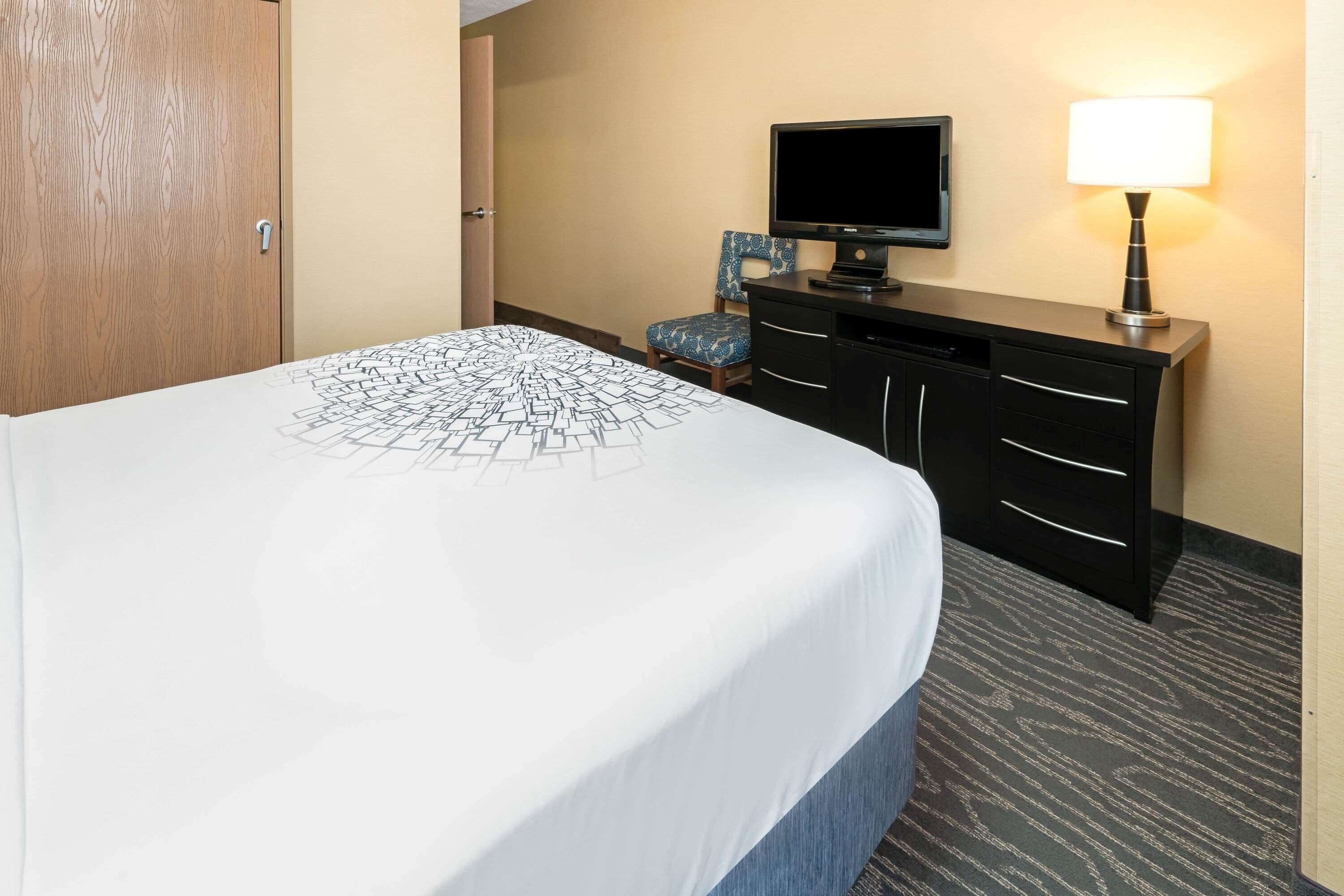 Una habitación de hotel con cama, televisión, escritorio con cajones, silla y lámpara.