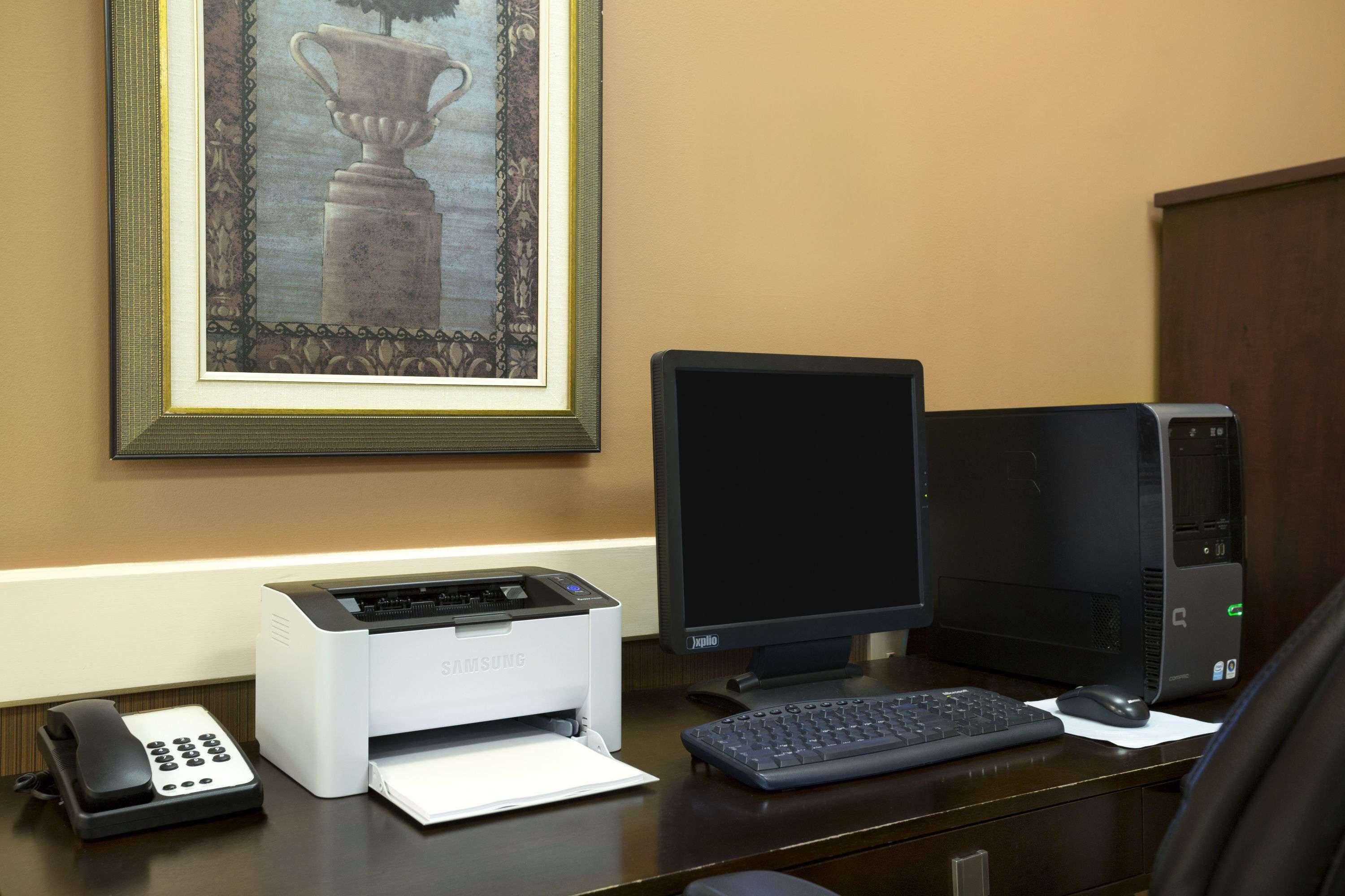 A desk with a desktop computer, a printer, a telephone, and a framed picture on the wall.
