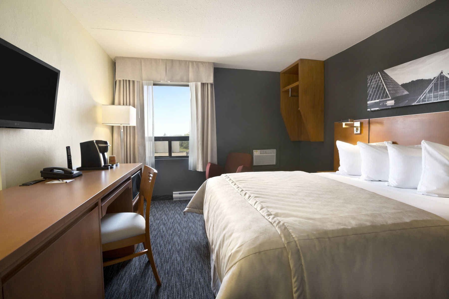 A hotel room with a bed, a desk with a telephone and lamp, a chair, a window with curtains, and a painting on the wall.