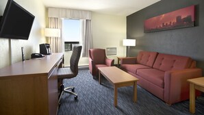 Una habitación de hotel con escritorio, sofá, televisor, lámpara y un cuadro de un paisaje urbano.