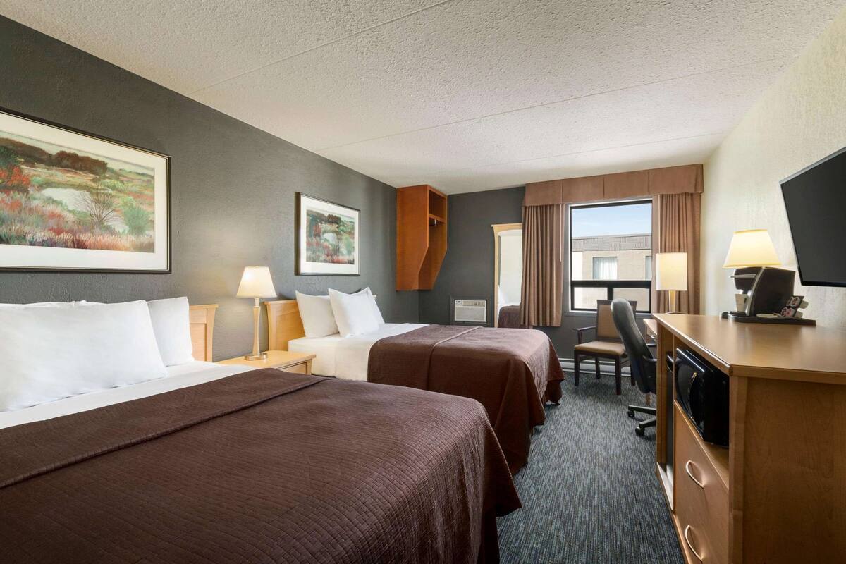 A hotel room with two beds, a desk with a lamp, a chair, and a large window.