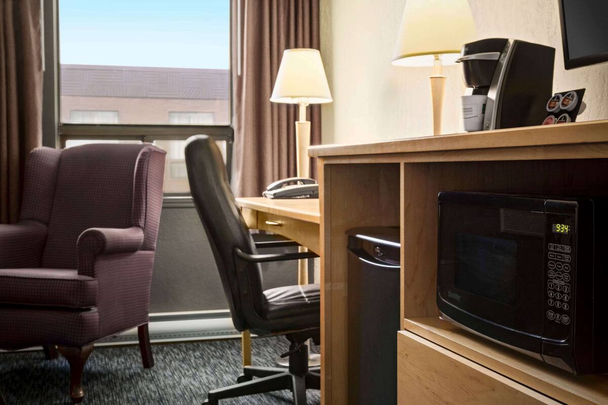 A hotel room with a desk, a microwave, a chair, a lamp, and a window with curtains.
