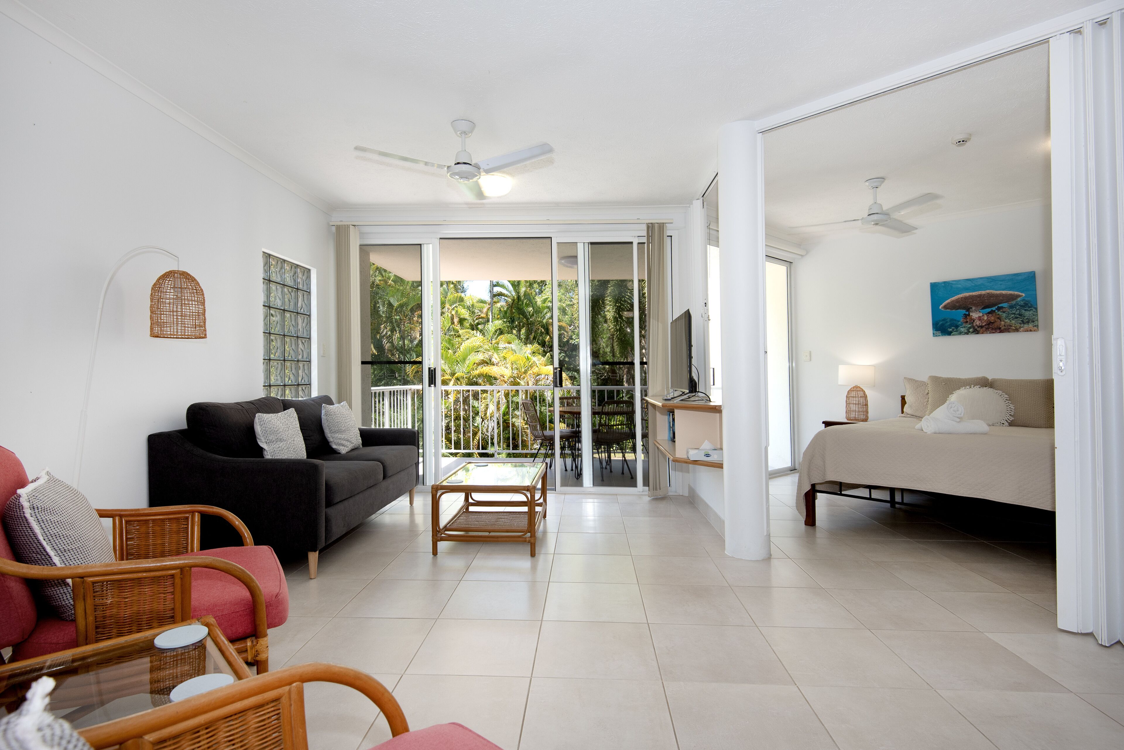A spacious living area with a black sofa, a wooden coffee table, wicker chairs, a bed, and a balcony with a view of palm trees.