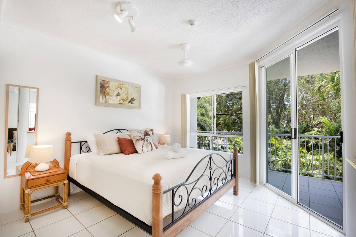 A bedroom with a bed, a nightstand, a mirror, a painting, and a balcony with a view of greenery.