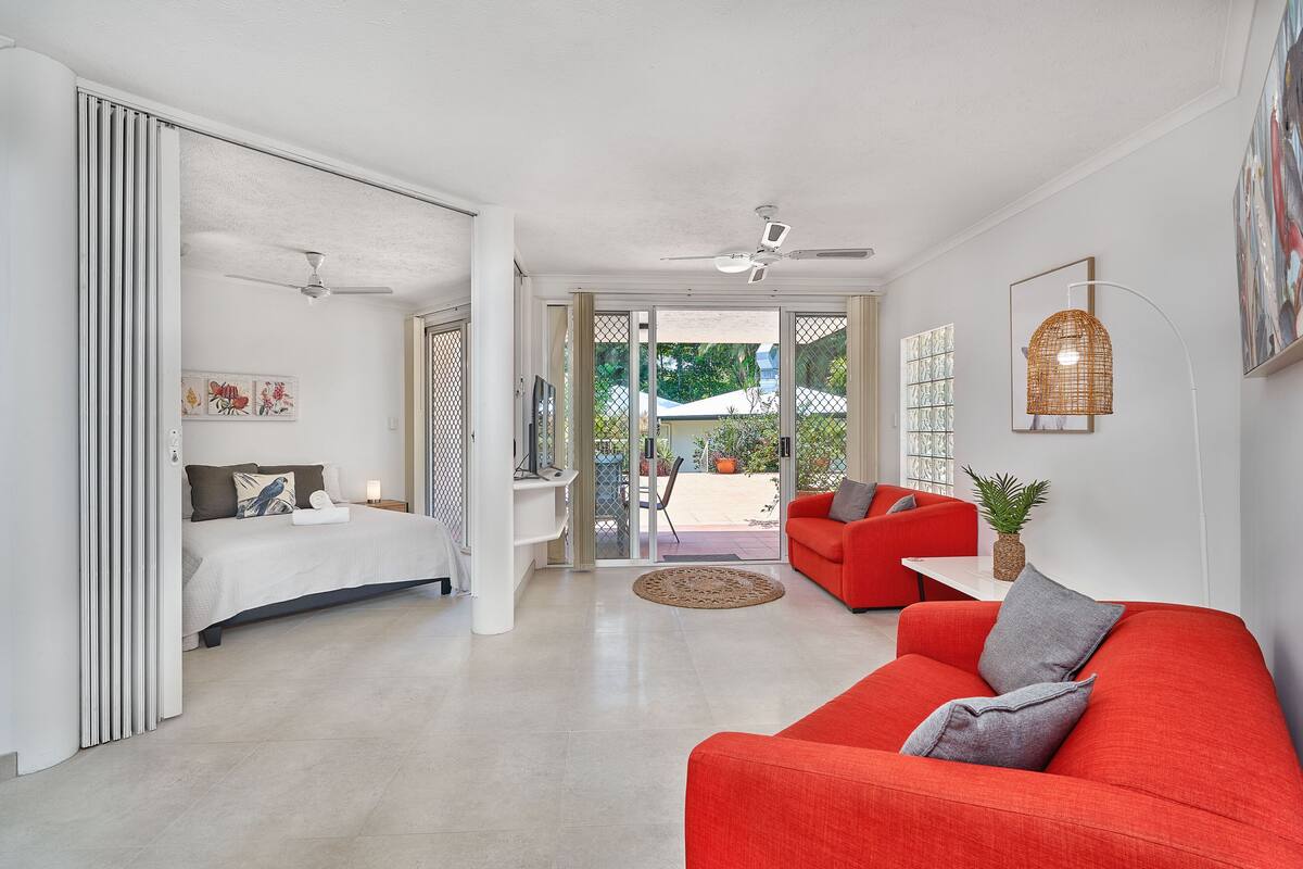 A modern living room with a red sofa, a bed with a white headboard, and a ceiling fan.