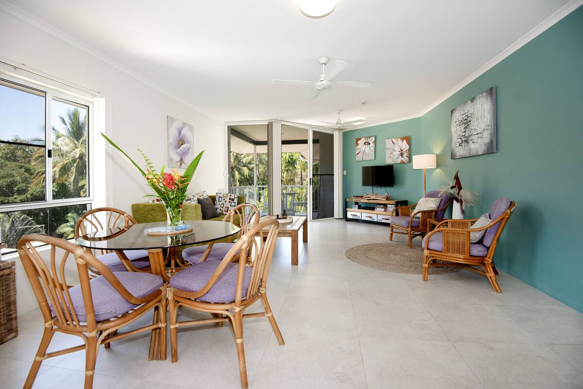 A spacious living area with a dining table surrounded by wicker chairs, a sofa, and a television.
