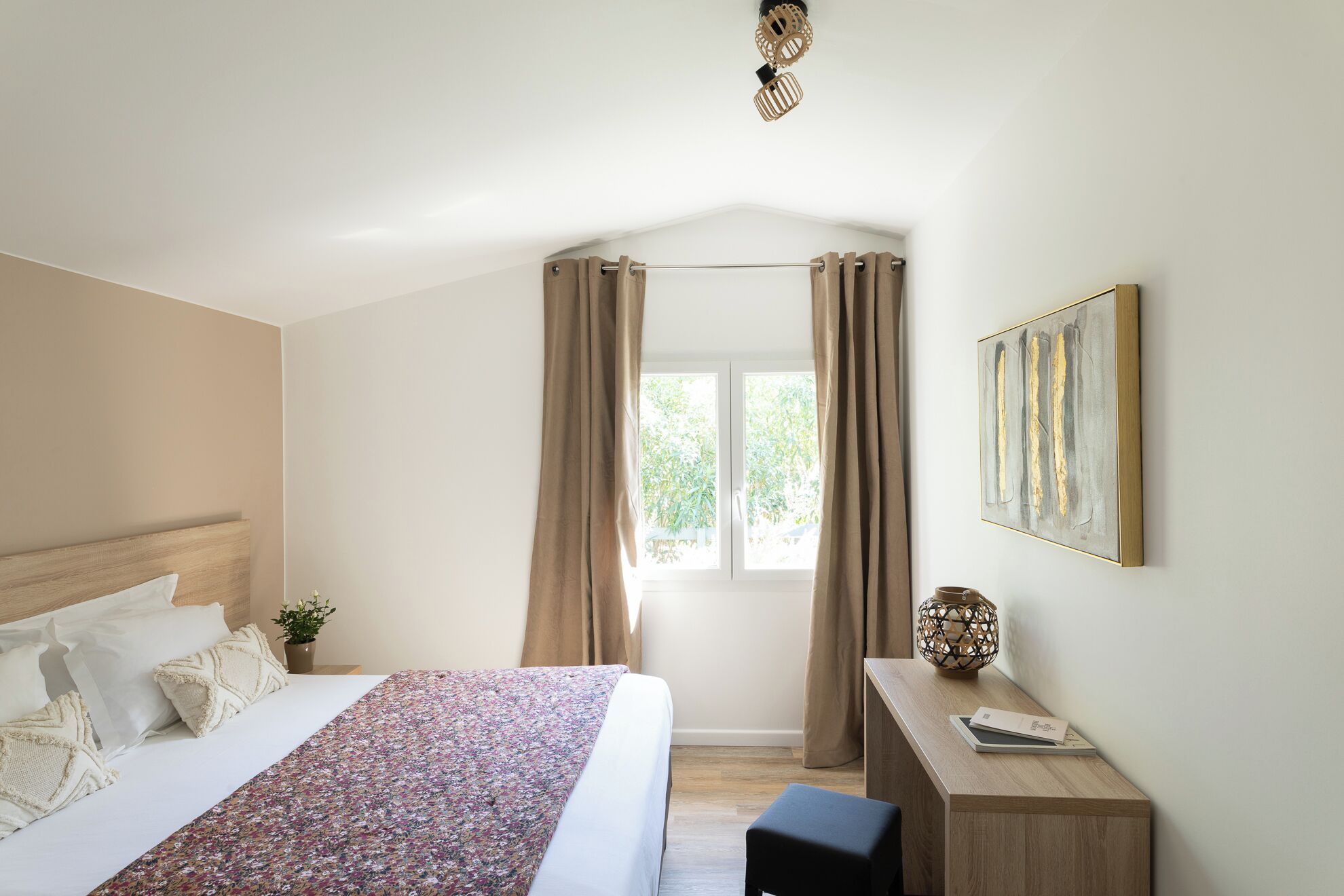 A hotel room with a bed, a desk, a window with curtains, and a painting on the wall.