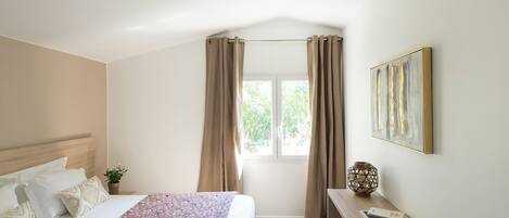 A hotel room with a bed, a desk, a window with curtains, and a painting on the wall.