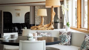 A modern lounge area with a round table, a sofa with floral cushions, and a unique wooden lamp stand. - Golfe Hôtel (Porto-Vecchio)
