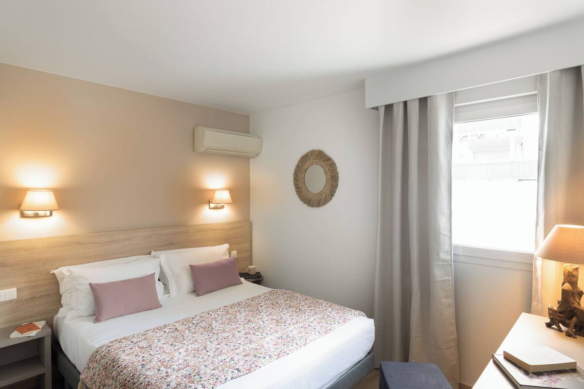Une chambre d’hôtel comprenant un lit, deux lampes fixées au mur, un appareil de climatisation, un miroir décoratif et une fenêtre avec des rideaux.