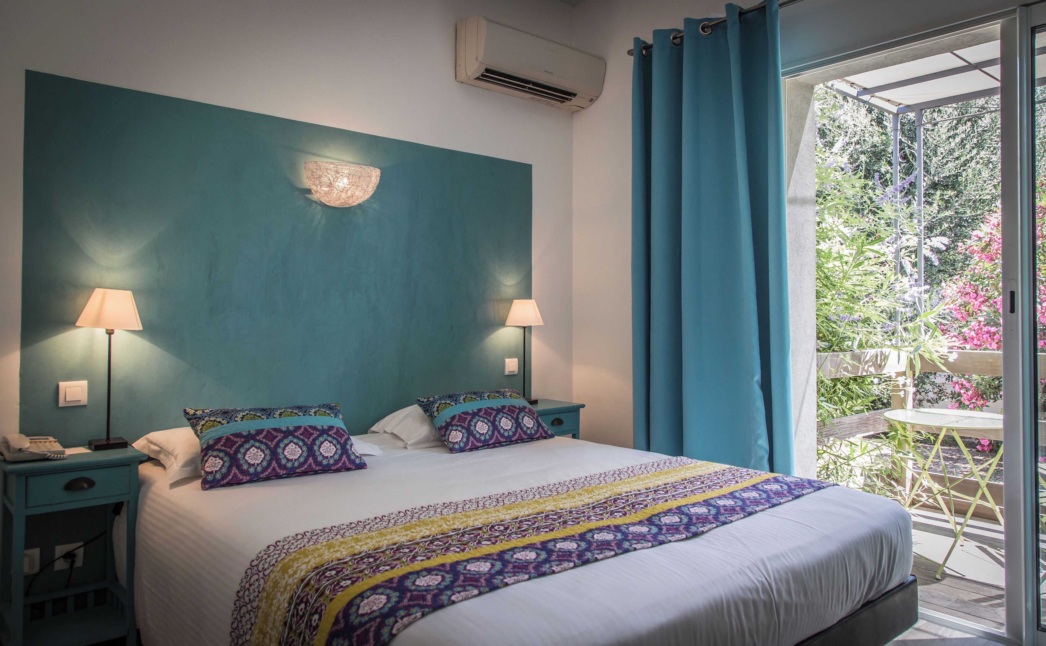 A bedroom with a large bed, two bedside lamps, a nightstand, and a view of a garden through a glass door.