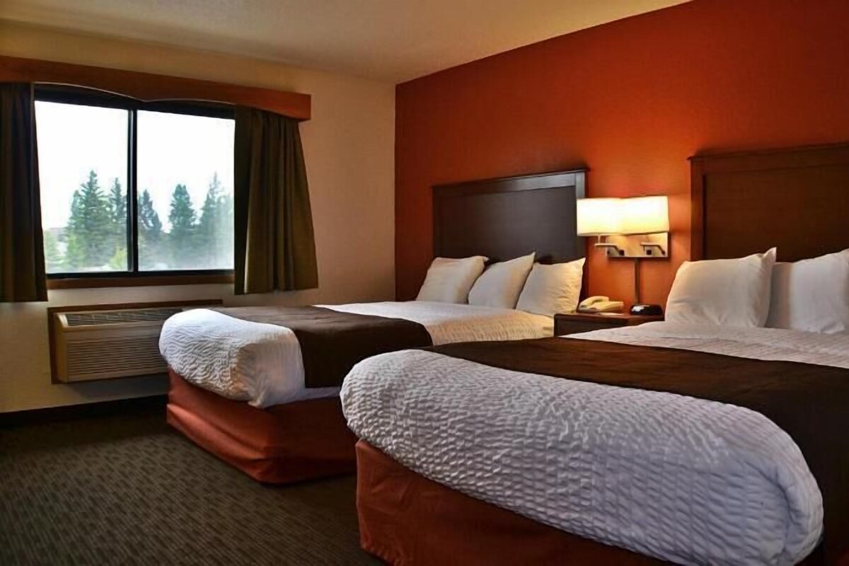 A hotel room with two beds, a window with curtains, a nightstand with a lamp, and a wall-mounted heater.