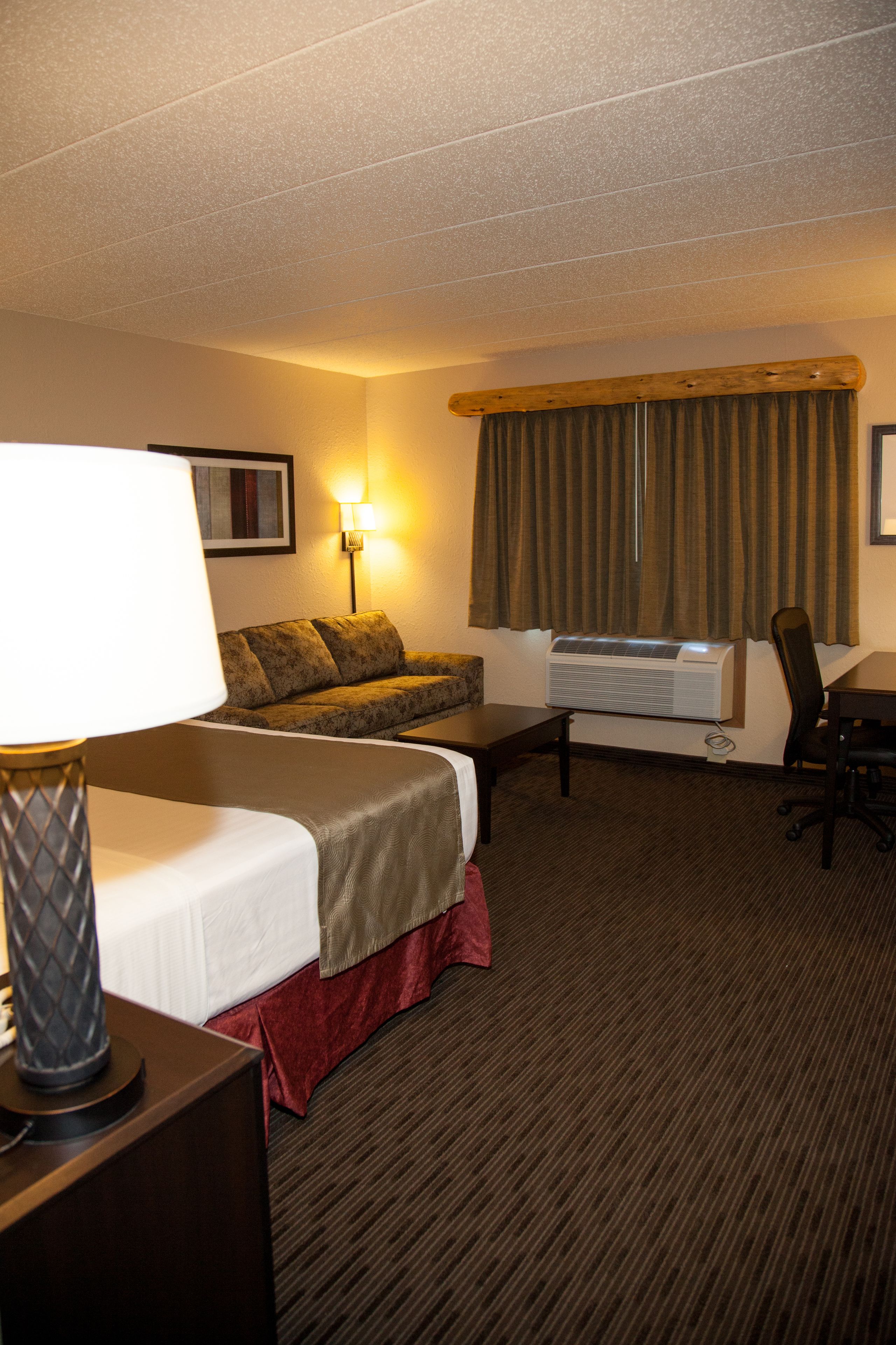 A hotel room with a bed, a desk, a chair, a lamp, a sofa, and an air conditioning unit.