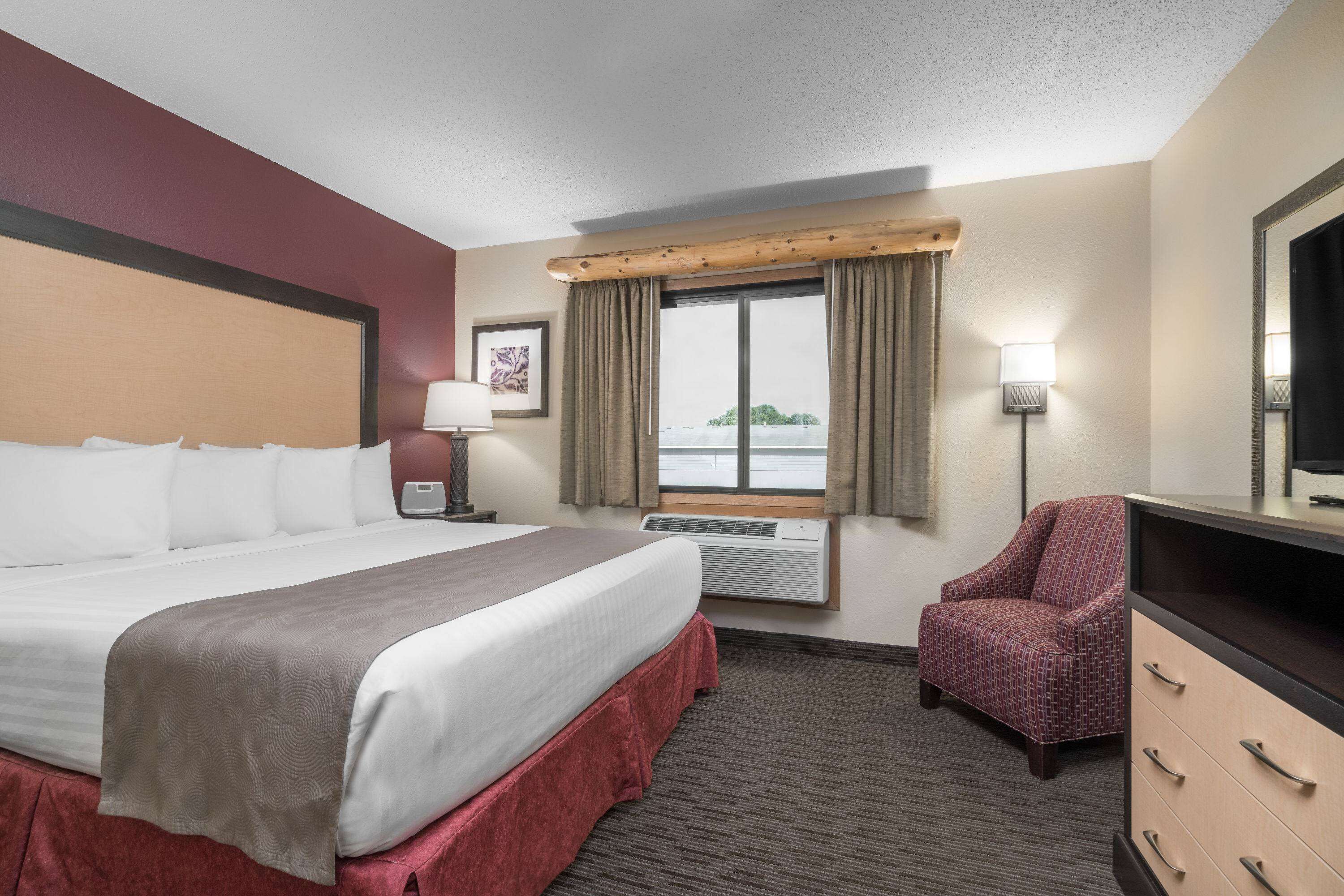 Quarto de hotel com uma cama grande, uma cadeira, uma escrivaninha e uma janela com cortinas.