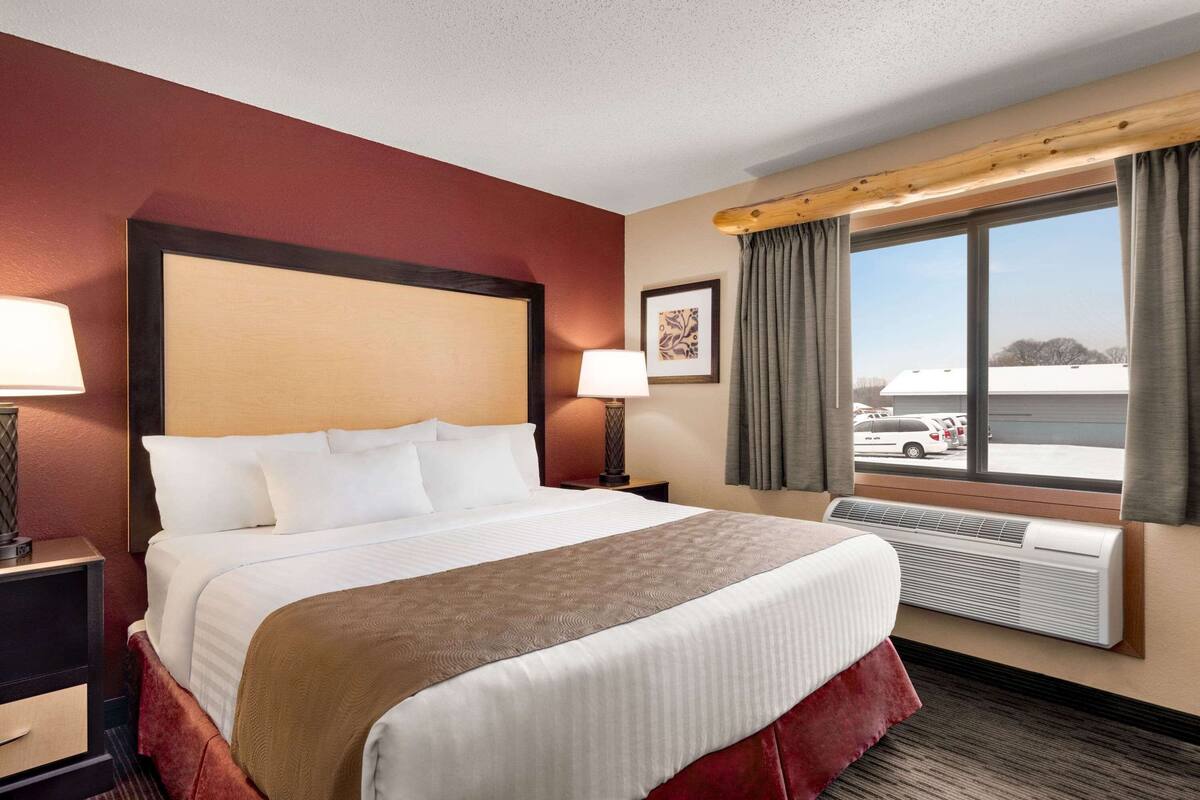 Ein Hotelzimmer mit einem großen Bett, einem Nachttisch, einer Lampe, einem Fenster mit Vorhängen und Blick nach draußen.