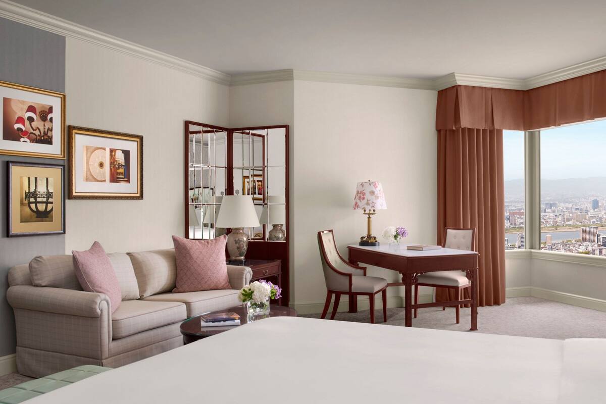 Habitación de hotel con una cama, un escritorio con una silla, un sofá, una mesa de centro y un ventanal con vistas a la ciudad.