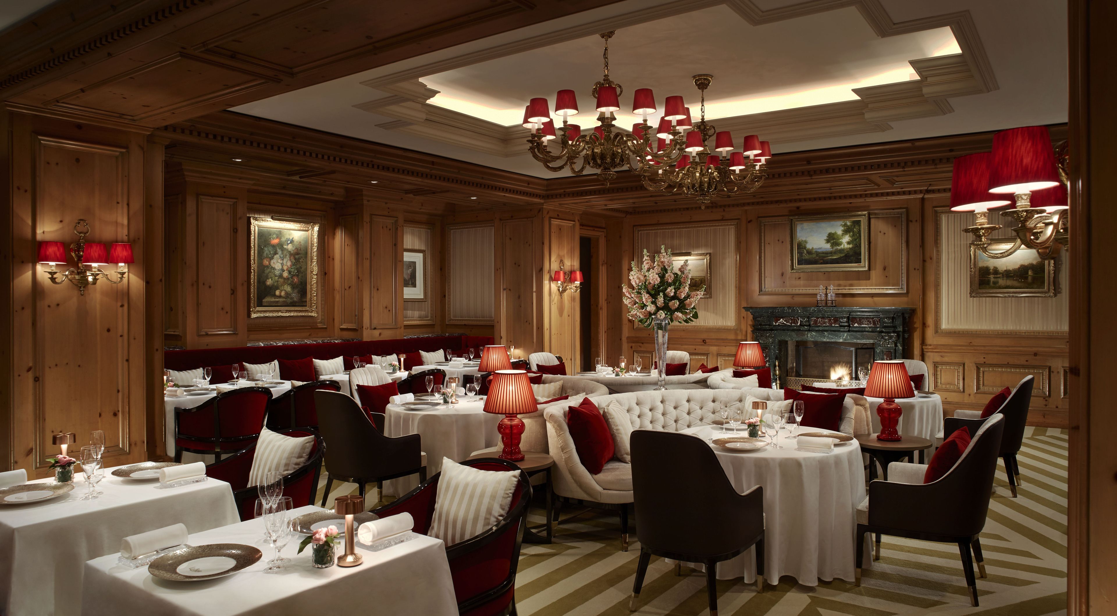 Uma sala de jantar formal com várias mesas, cadeiras estofadas em vermelho e um candelabro.