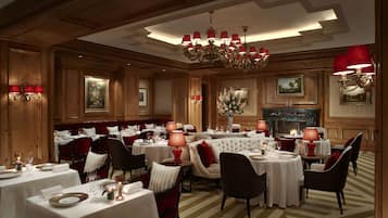 Uma sala de jantar formal com várias mesas, cadeiras estofadas em vermelho e um candelabro.