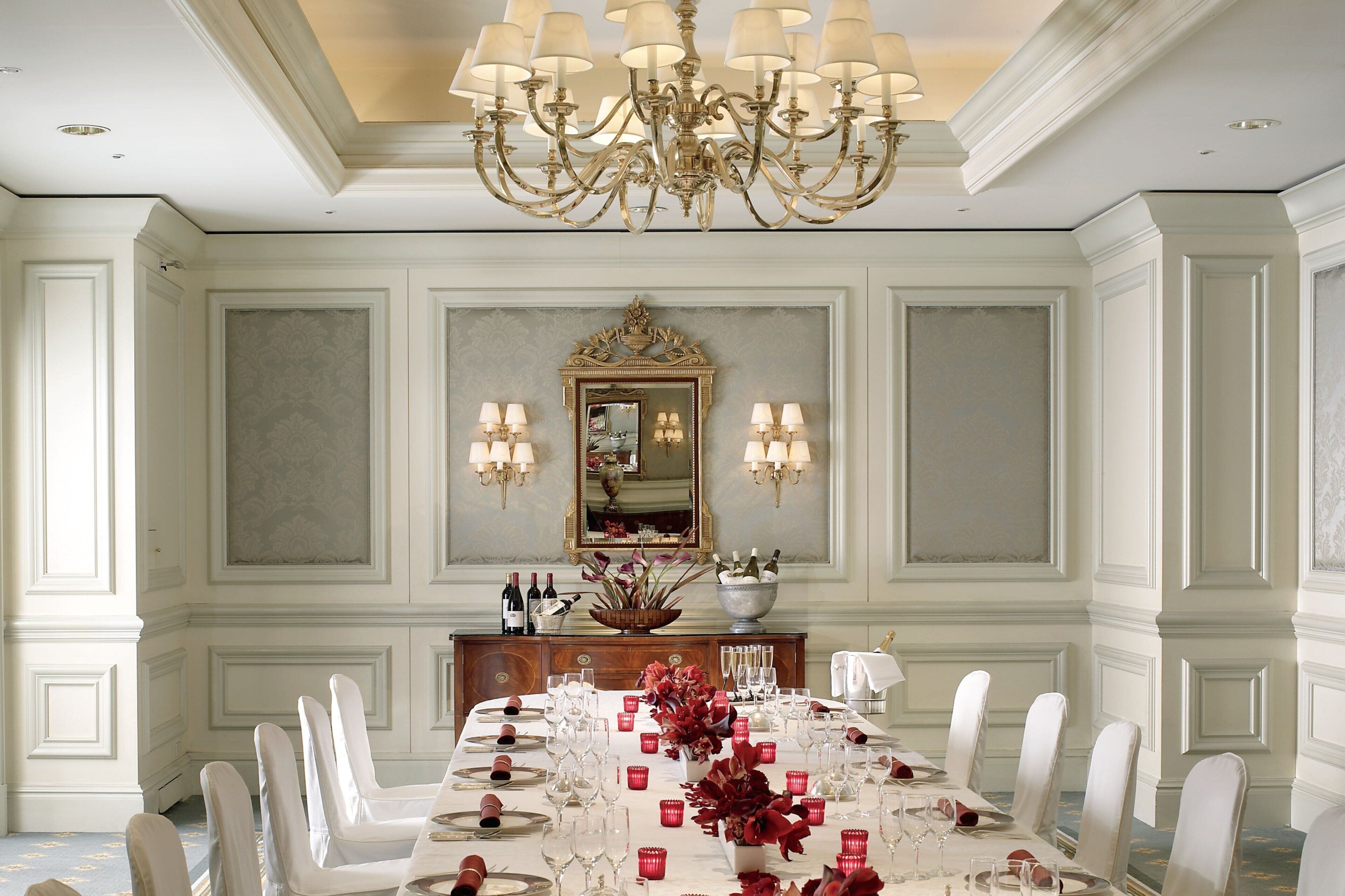 A formal dining room with a long table set for a meal, elegant chairs, a chandelier, and a decorative mirror above the table.