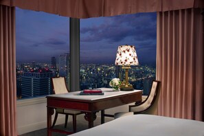 A hotel room with a desk, chair, and a lamp. There is a view of a cityscape through large windows.