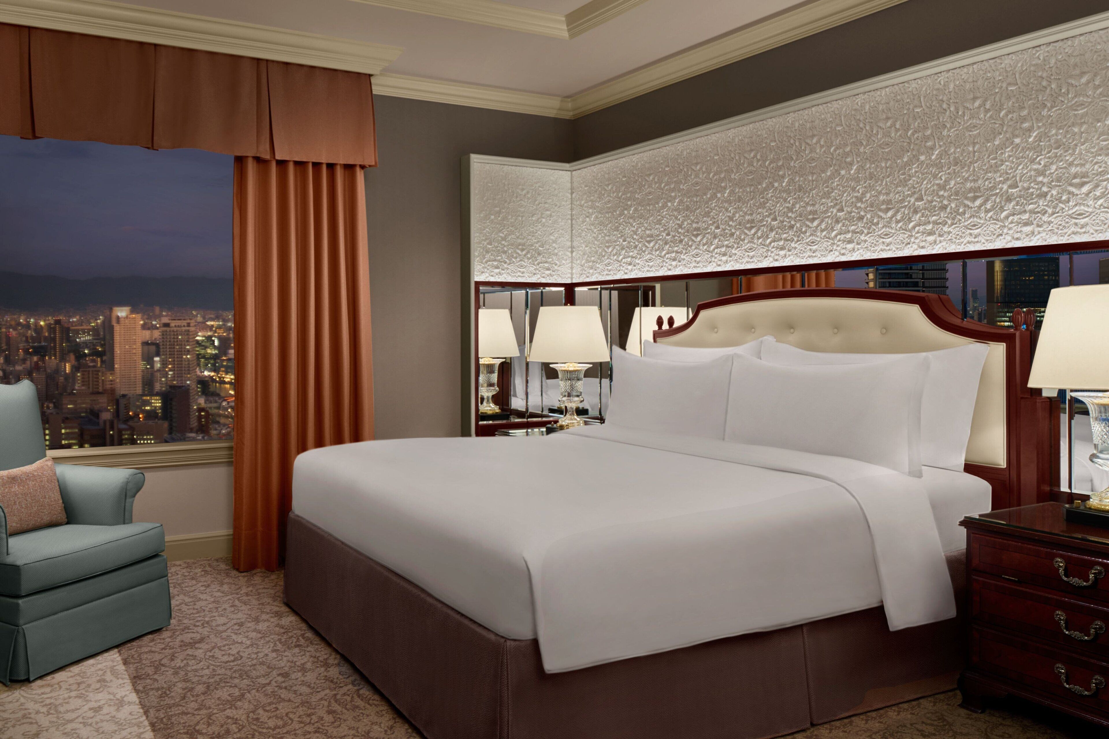 A hotel room with a large bed, a nightstand, a chair, and a view of a cityscape.