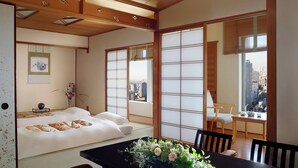 A traditional Japanese room with tatami flooring, a low table with a vase of flowers, a bed with white bedding, and large windows offering a city view. - The Ritz-Carlton, Osaka (Osaka)