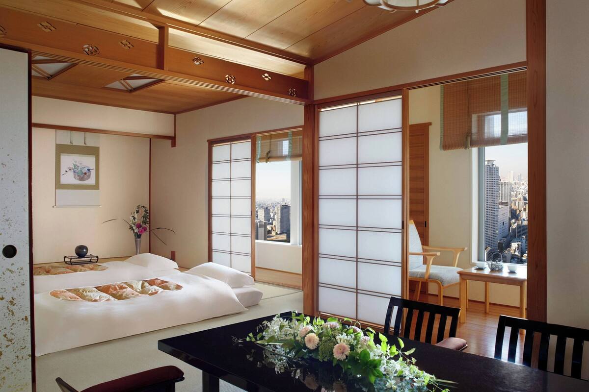 Una habitación japonesa tradicional con suelo de tatami, una mesa baja con un jarrón de flores, una cama con ropa de cama blanca y grandes ventanales con vistas a la ciudad.