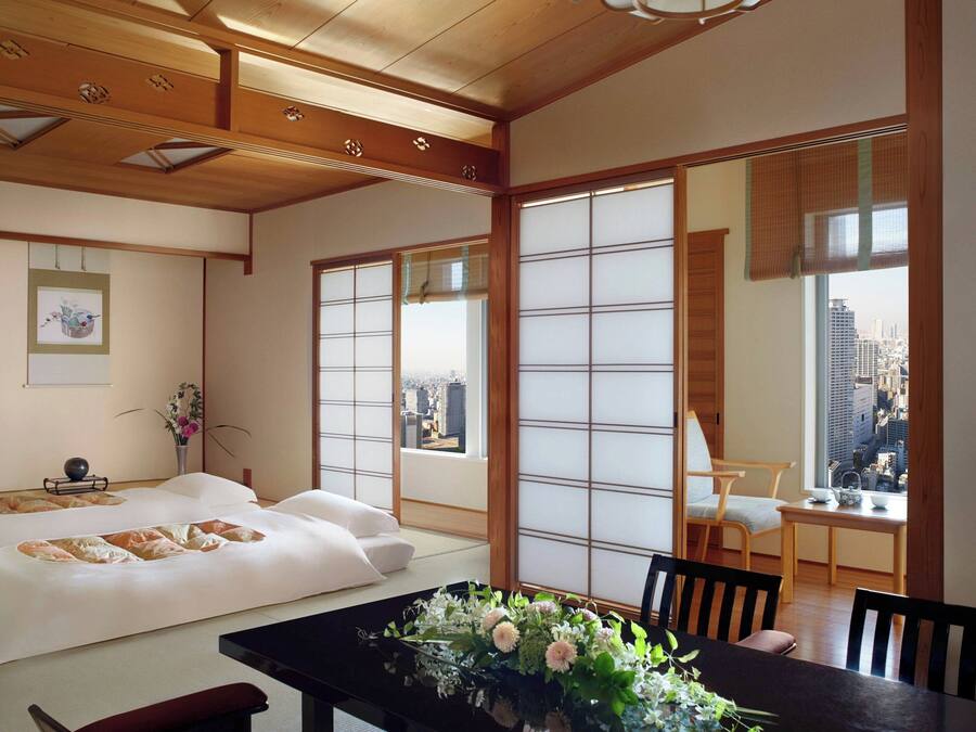 Una habitación japonesa tradicional con suelo de tatami, una mesa baja con un jarrón de flores, una cama con ropa de cama blanca y grandes ventanales con vistas a la ciudad.