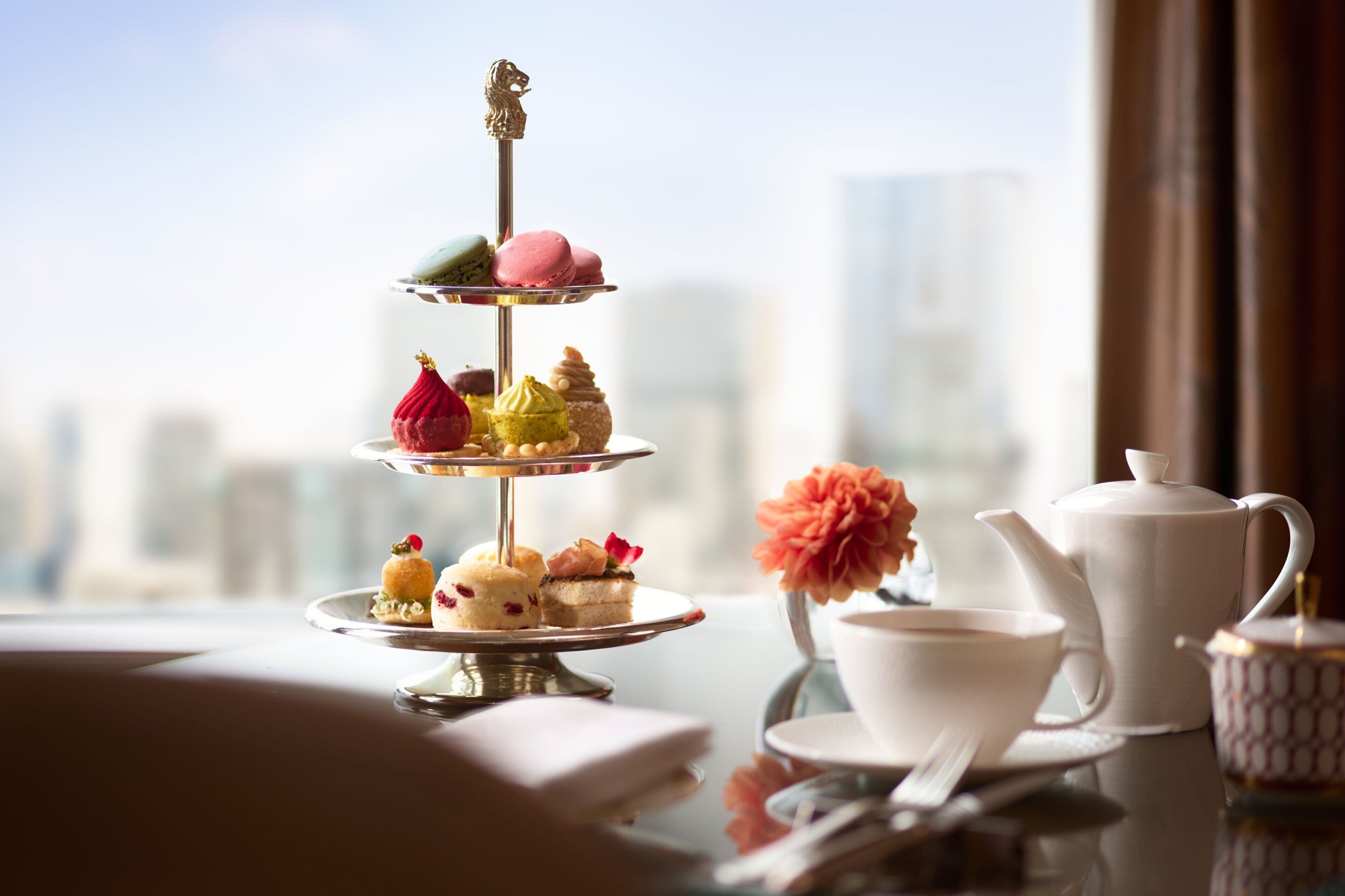 Un expositor escalonado para postres con diversas pastelerías y una tetera sobre una mesa con vista a un paisaje urbano.