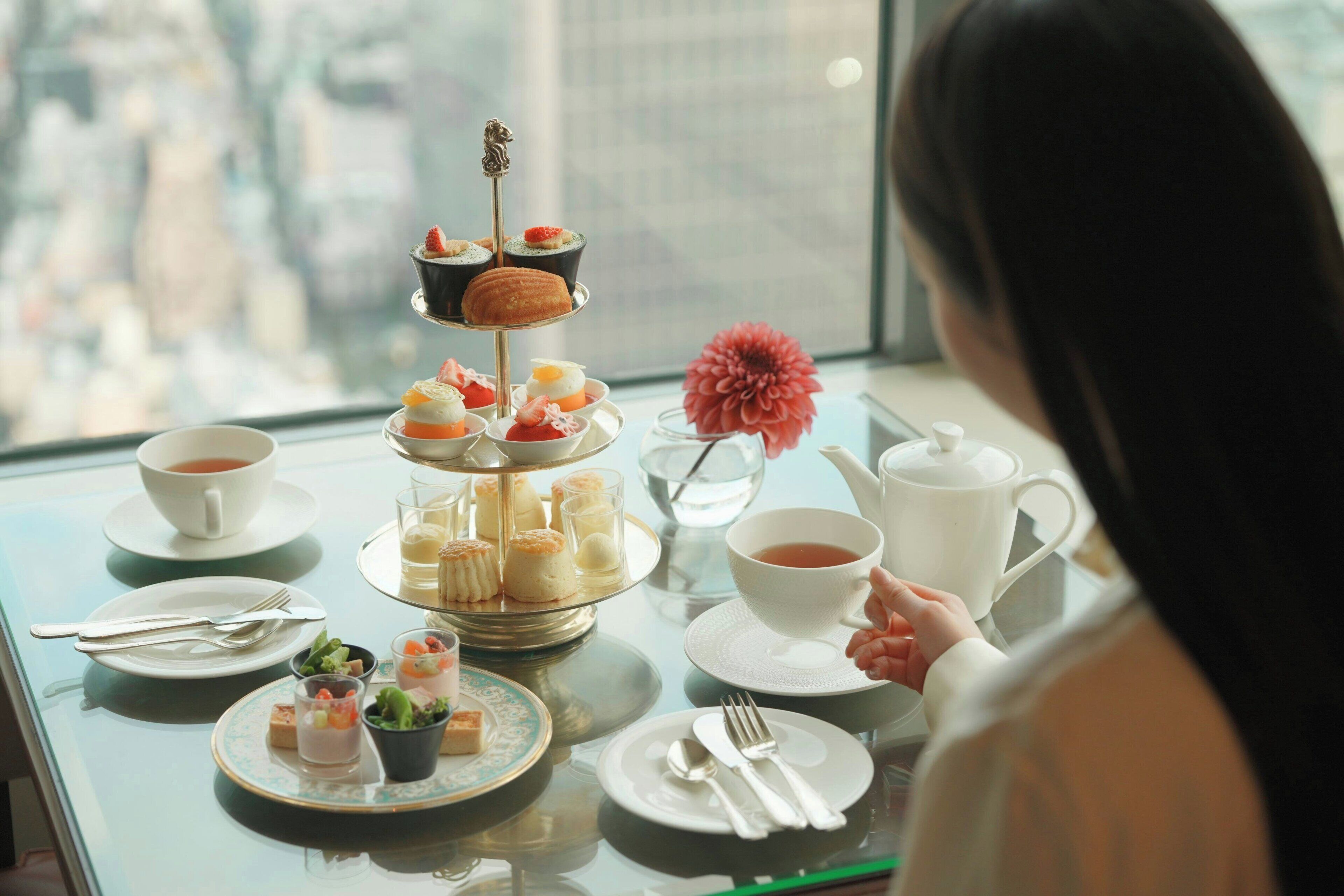 A table set for a tea service with various pastries and desserts, a flower vase, and a city view.