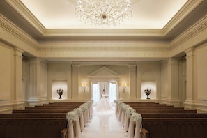 A grand hall with a central aisle, flanked by rows of wooden pews, leading to an arched doorway.