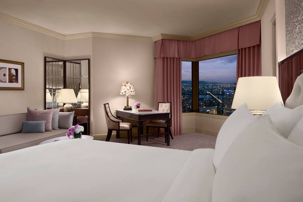 Habitación de hotel con una cama grande, un escritorio con dos sillas, vistas de la ciudad a través de la ventana y un sofá con cojines.