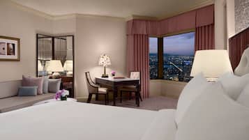 A hotel room with a large bed, a desk with two chairs, a city view through the window, and a sofa with pillows.