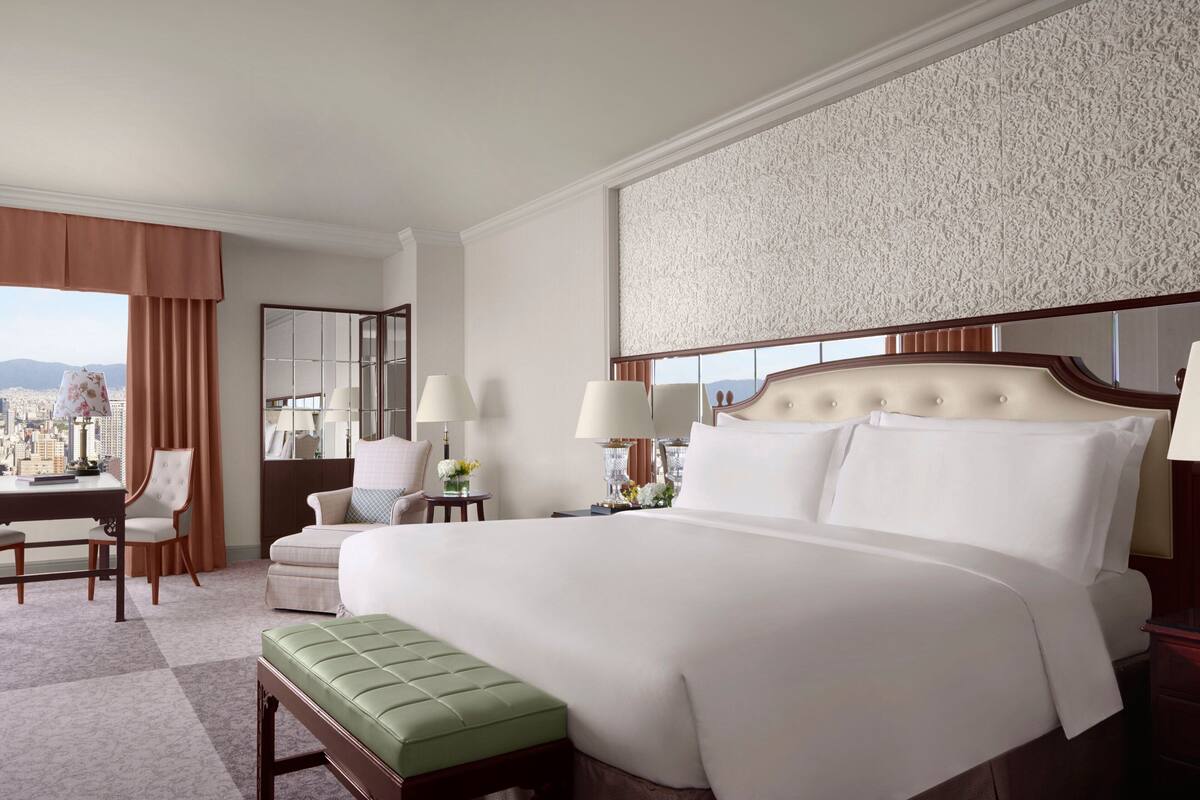 Habitación de hotel con una cama grande, un banco verde, un escritorio con una silla y vistas a la ciudad.