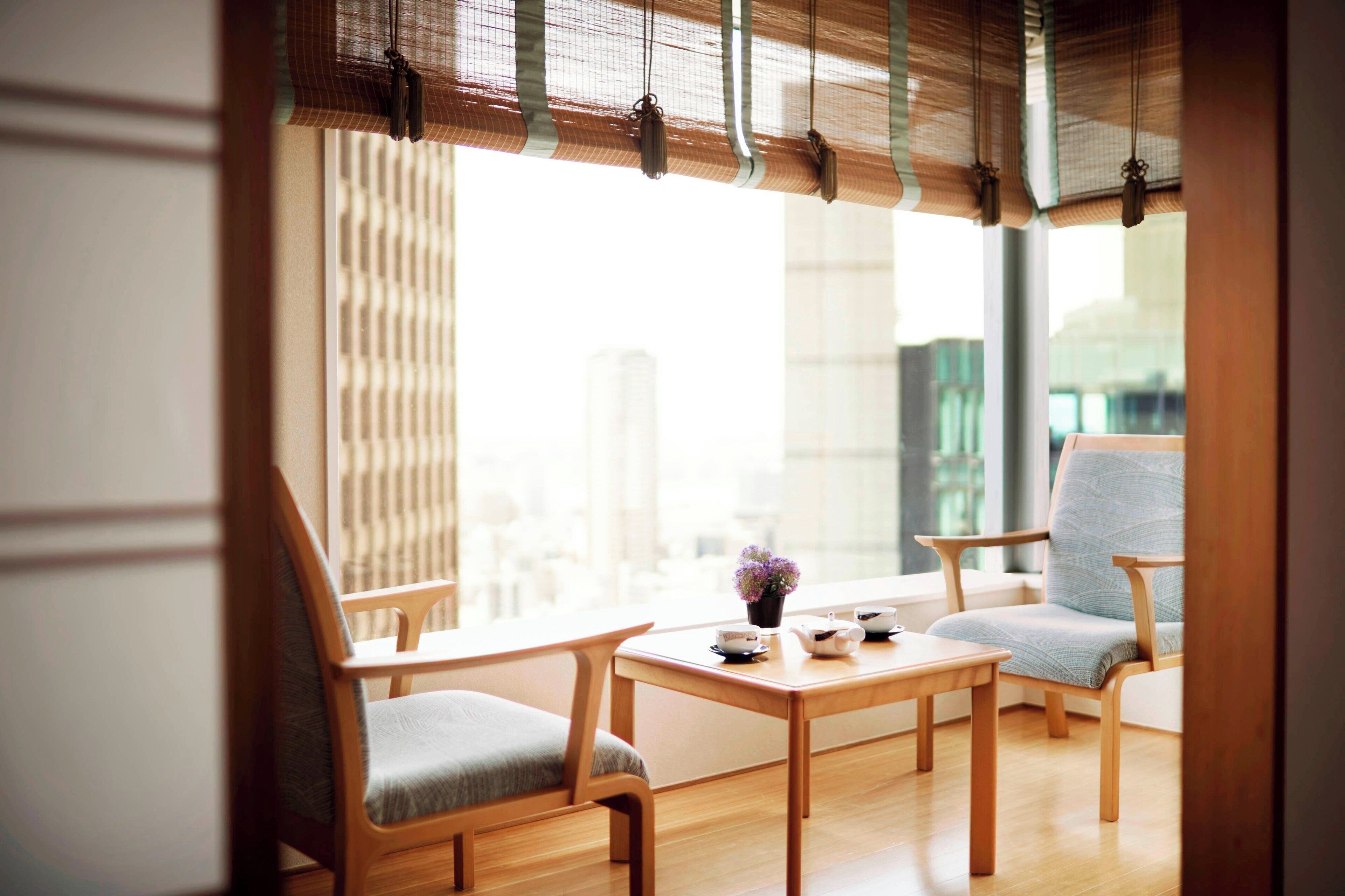 Una habitación con piso de madera, dos sillas de madera, una mesita y una ventana con vistas a edificios.