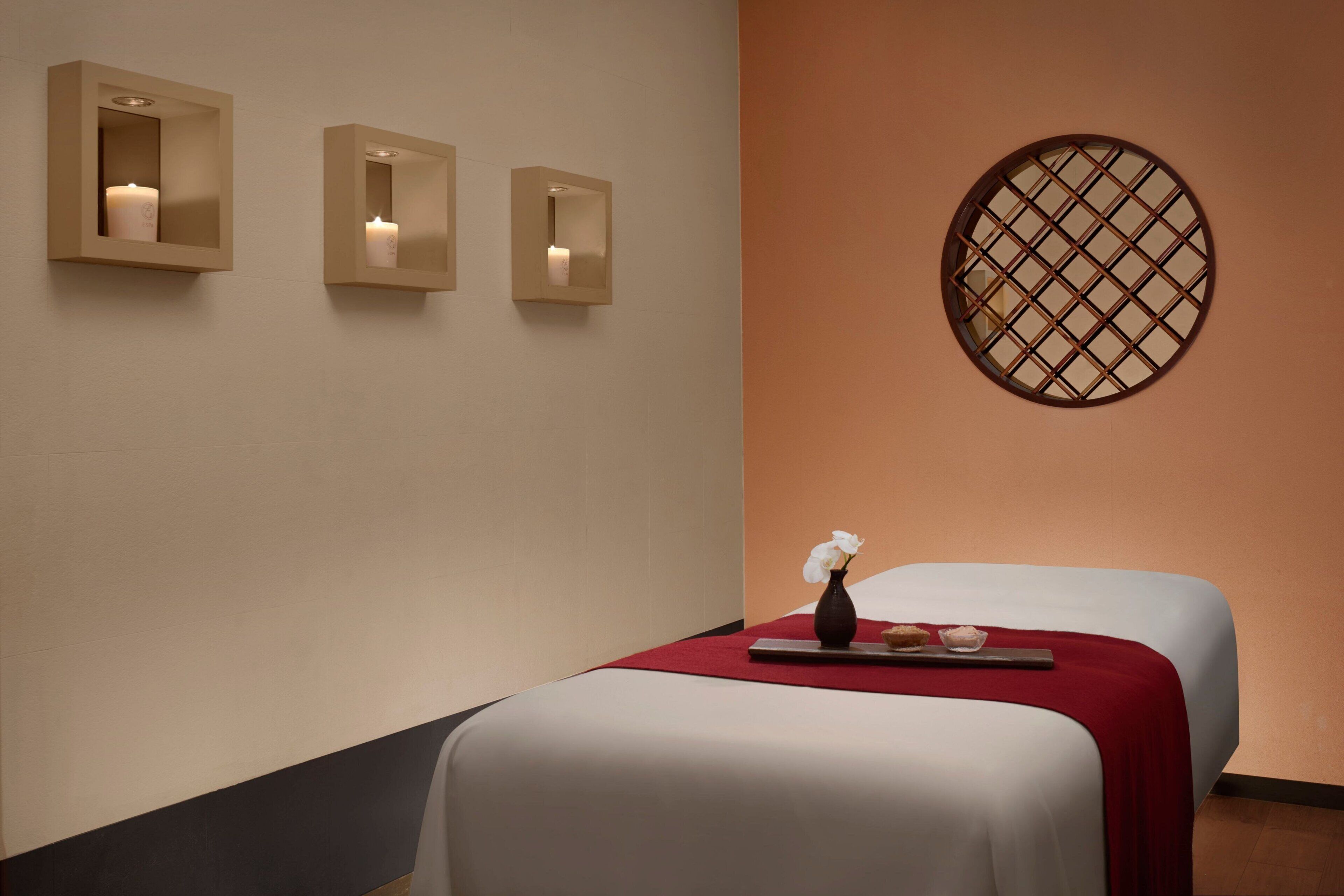 A hotel room with two beds, a red throw blanket, a vase with a flower, and three wall-mounted sconces with candles.