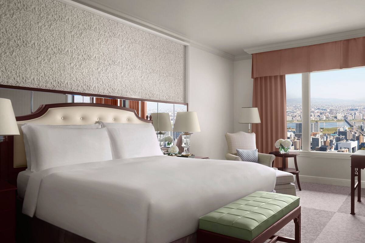 Habitación de hotel con una cama grande, dos mesitas de noche con lámparas, un banco verde y vistas a un paisaje urbano a través de amplias ventanas.