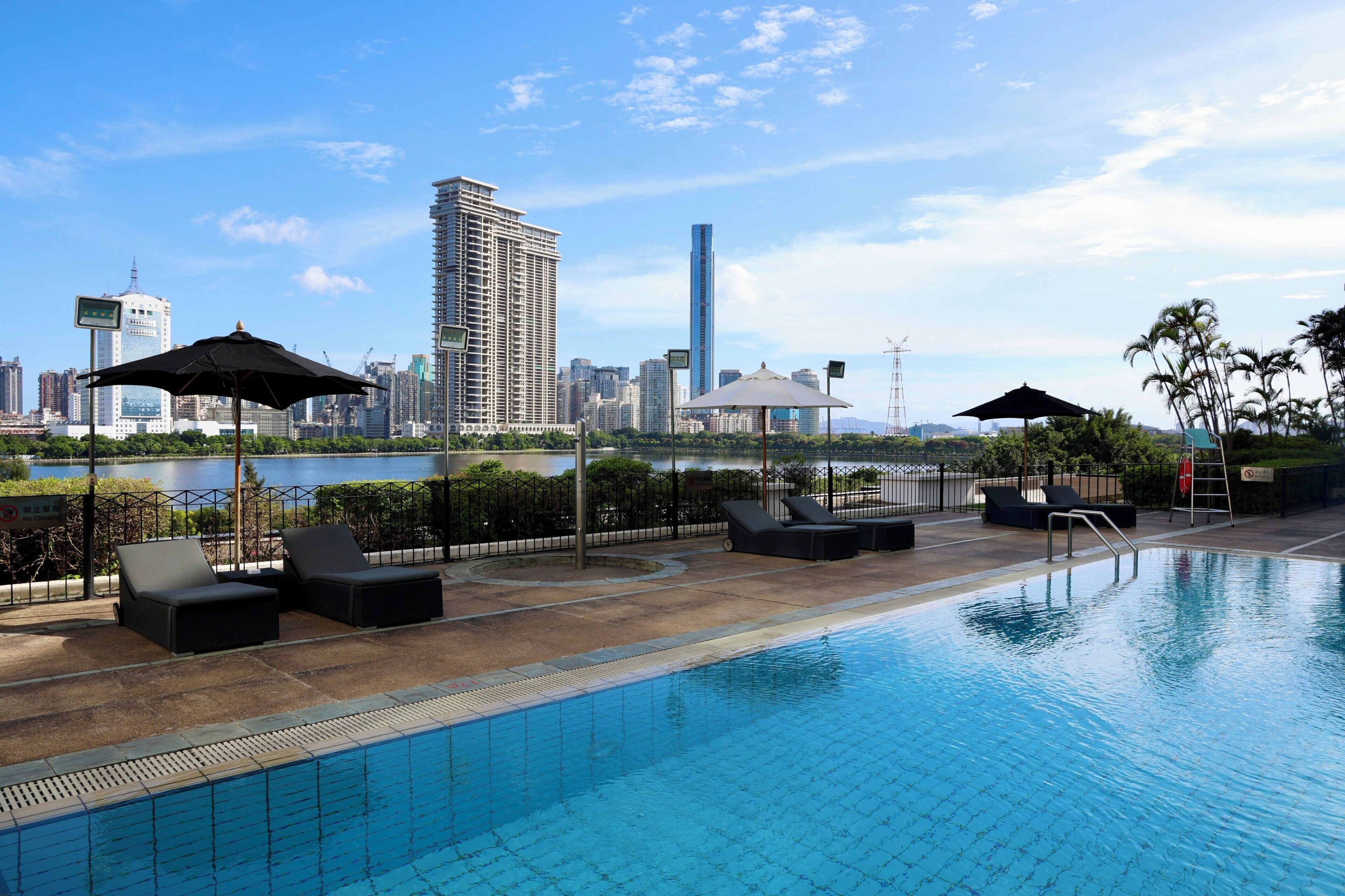 A swimming pool with lounge chairs and umbrellas, a view of a city skyline, and a body of water in the background.
