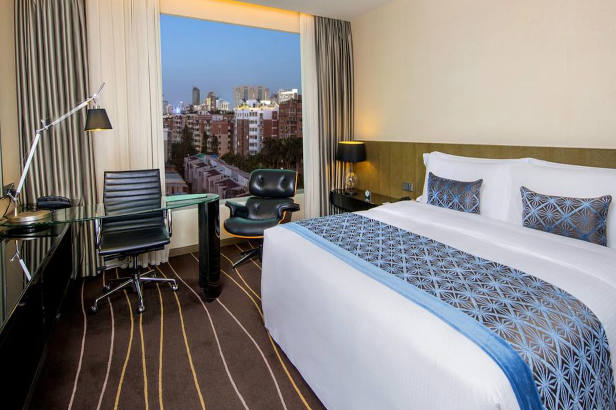 A hotel room with a large bed, a desk, a chair, a desk lamp, and a view of the cityscape.
