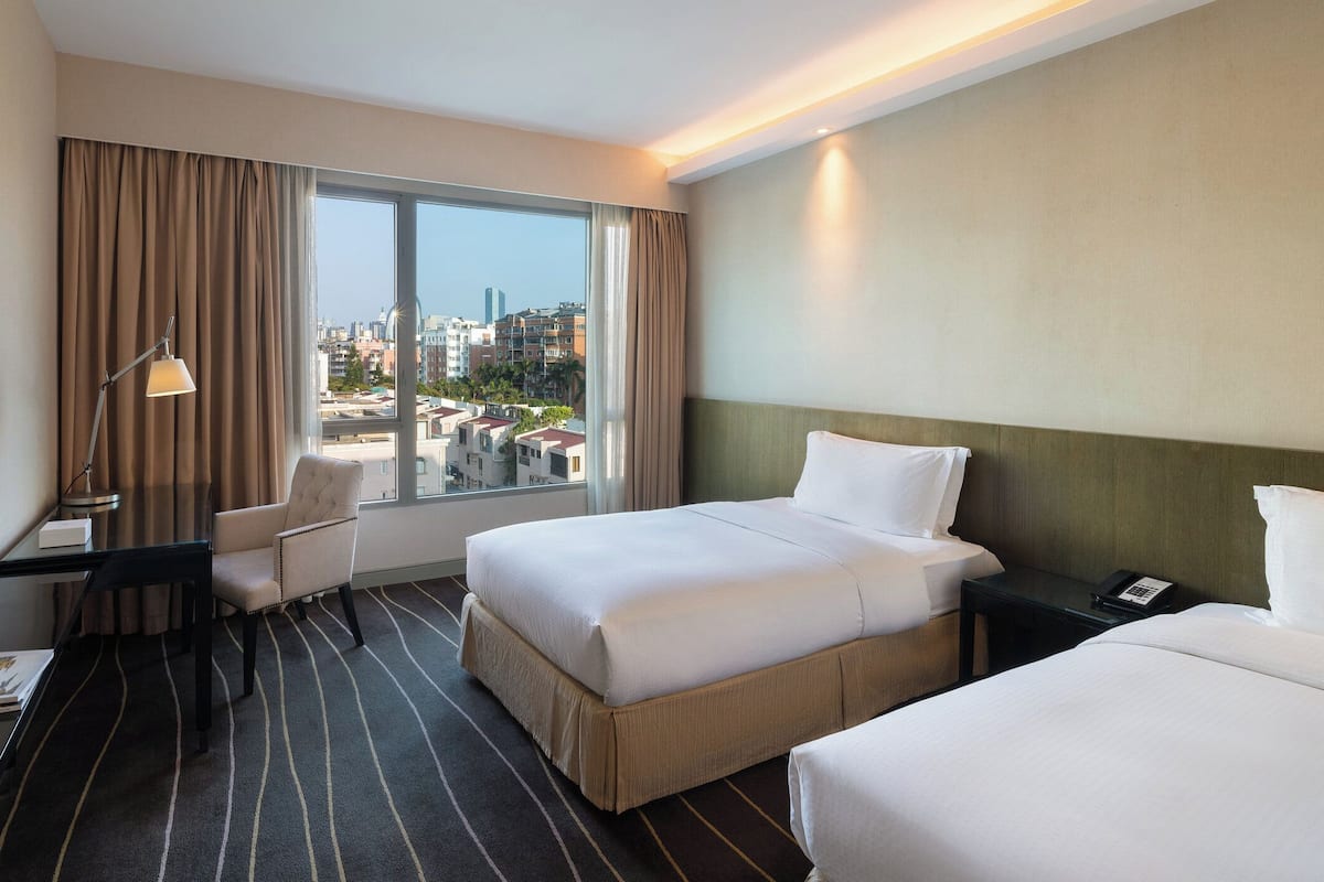 A hotel room with a large bed, a desk, a chair, and a view of a cityscape through a large window.