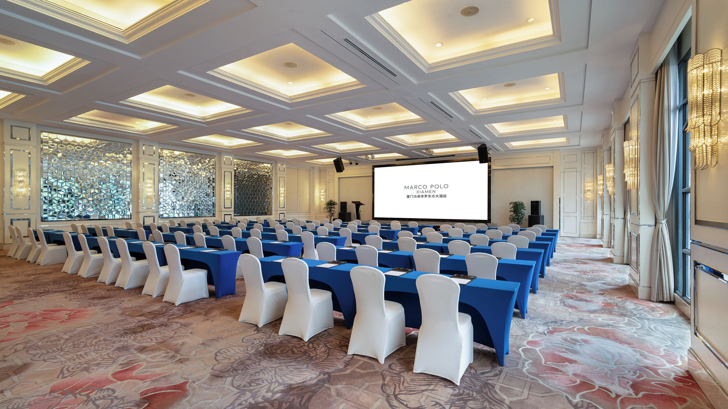 A conference room with rows of tables and chairs, a large screen, and a patterned carpet.