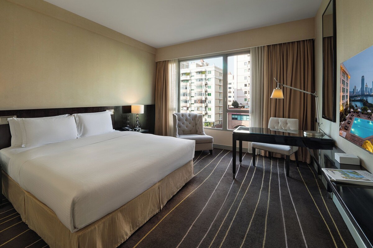 A hotel room with a large bed, a desk, a chair, a view of a cityscape, and a pool area.