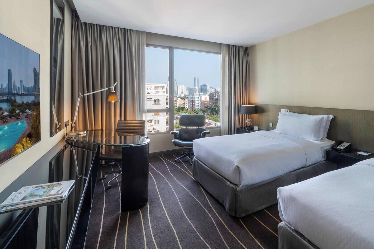 A hotel room with two beds, a desk, a chair, a view of the city, and a pool.