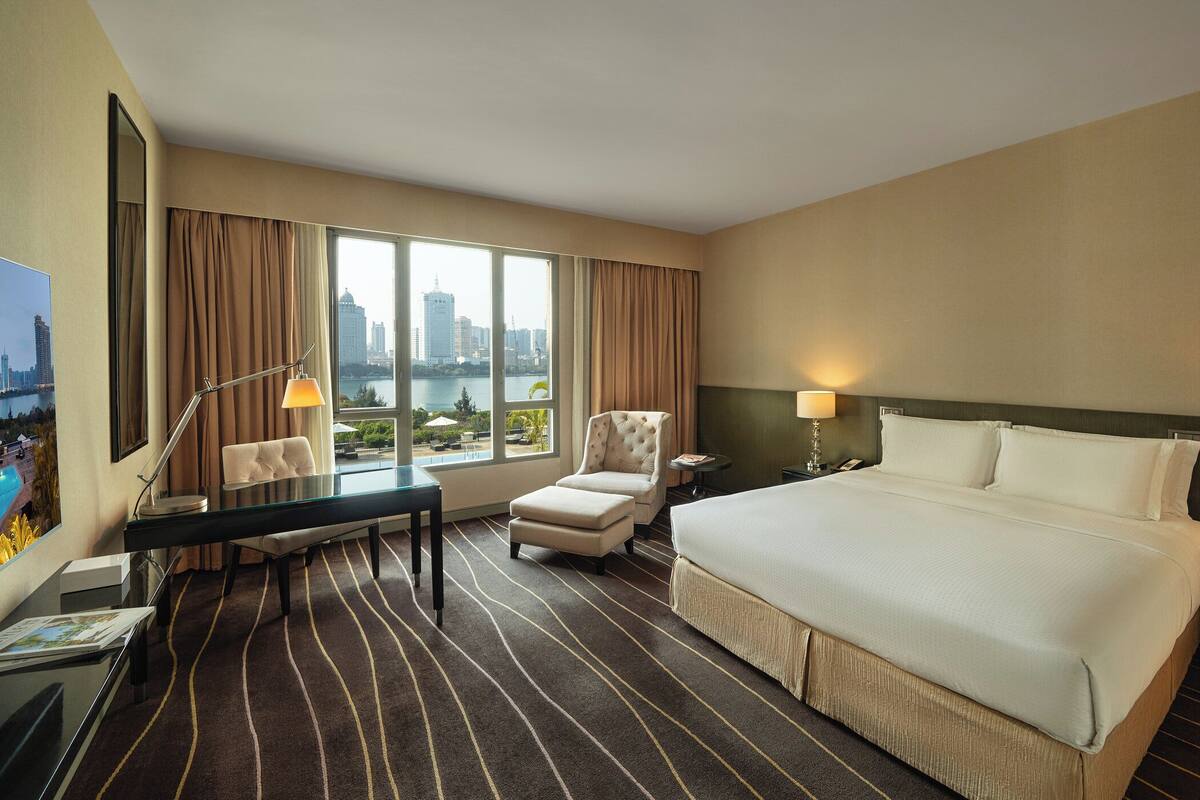 A hotel room with a large bed, a desk with a chair, a small table, and a view of a cityscape through large windows.
