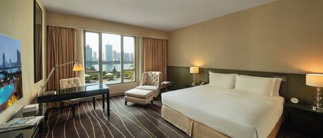 A hotel room with a large bed, a desk with a chair, a small table, and a view of a cityscape through large windows.