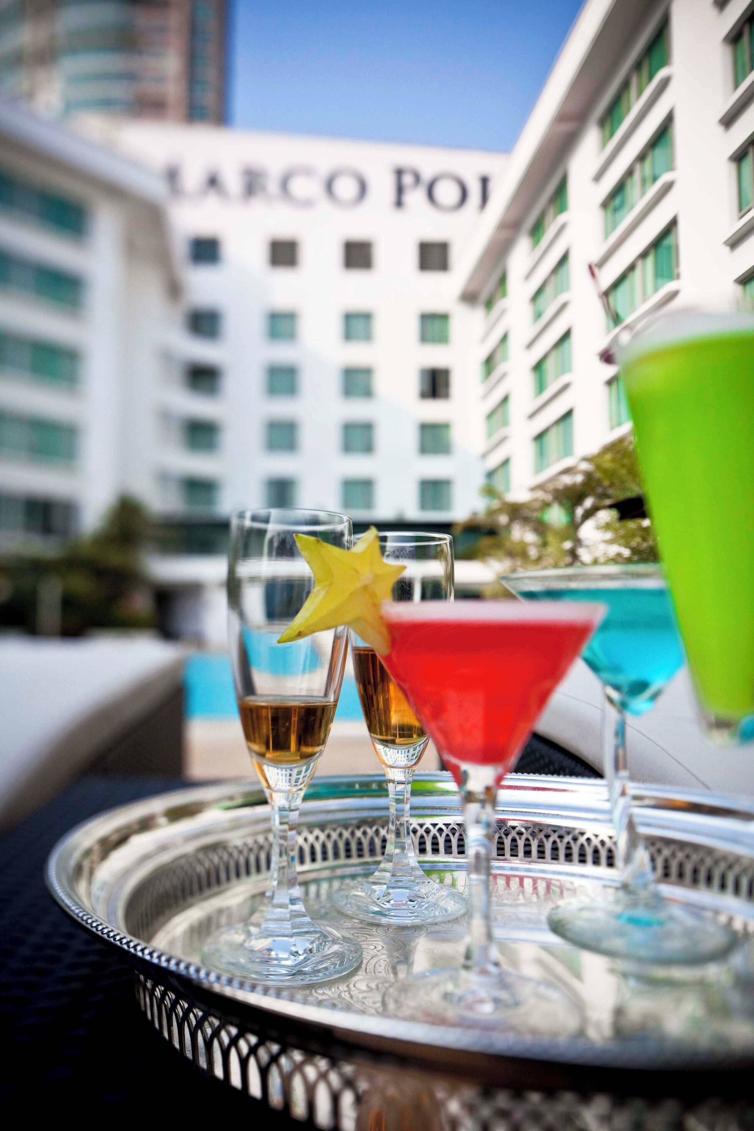 A tray with three glasses of beverages, one with a yellow star, and a green cocktail, set against a hotel backdrop with "MARCO POJ" signage.