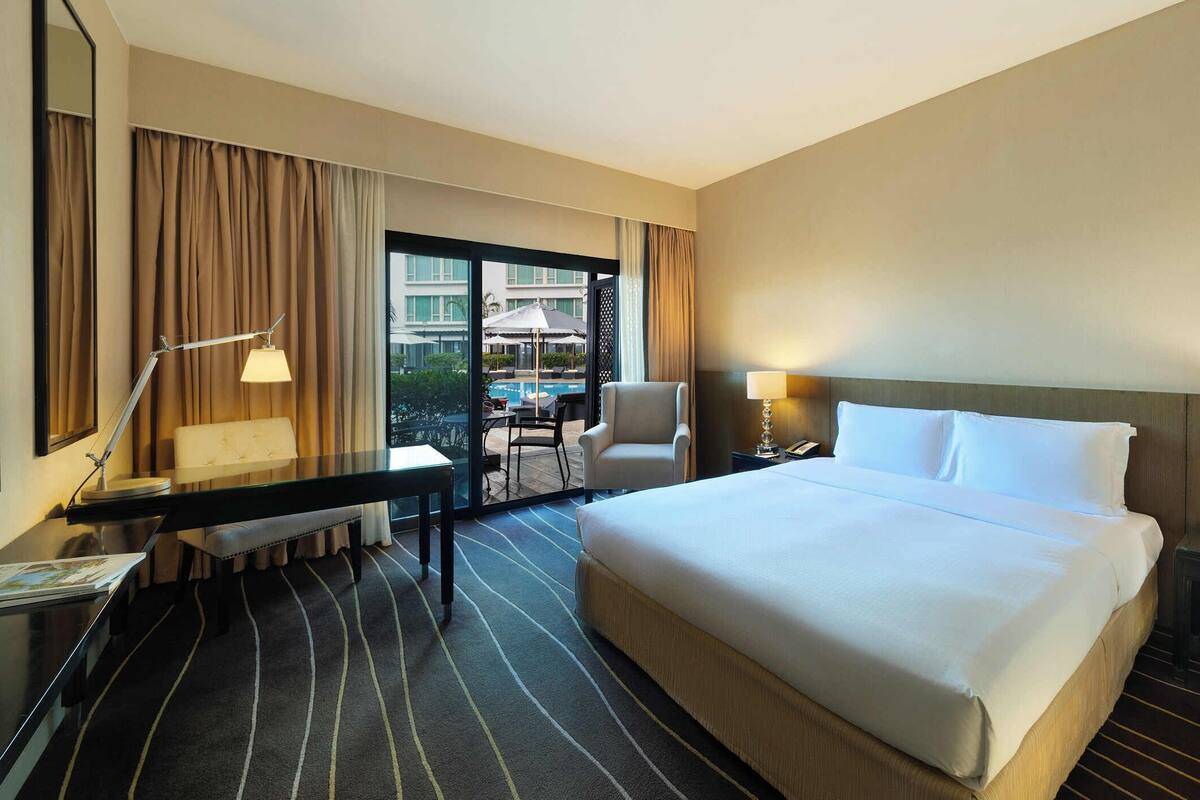 A hotel room with a large bed, a desk with a chair, a desk lamp, a nightstand with a lamp, and a view of an outdoor pool area.