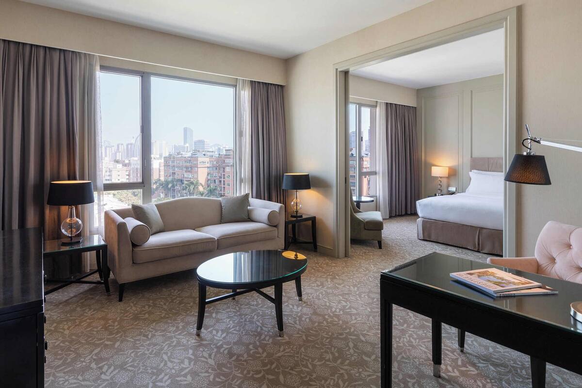 A hotel room with a sofa, a small table, a bed, and a view of the cityscape through large windows.
