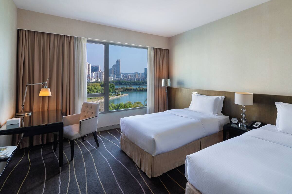 A hotel room with two beds, a desk, a chair, and a view of a cityscape and river.